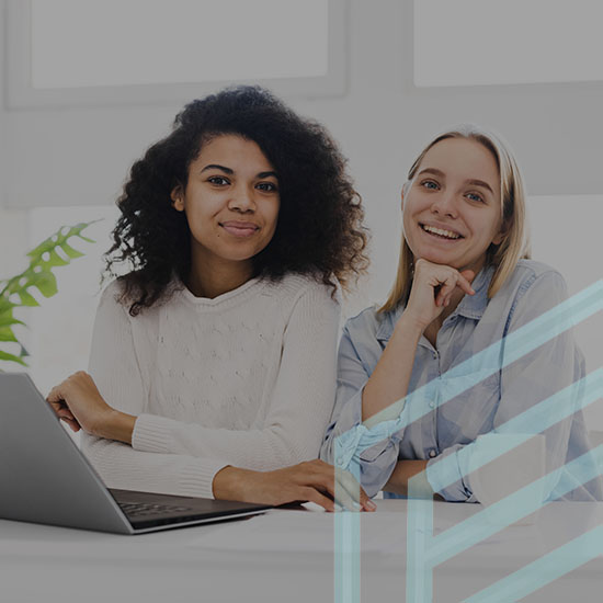 Two women sit at a desk with a laptop. One has curly hair and is wearing a white sweater, while the other has straight hair and is wearing a checkered shirt. They both smile towards the camera. A plant is in the background next to a window, adding natural light.