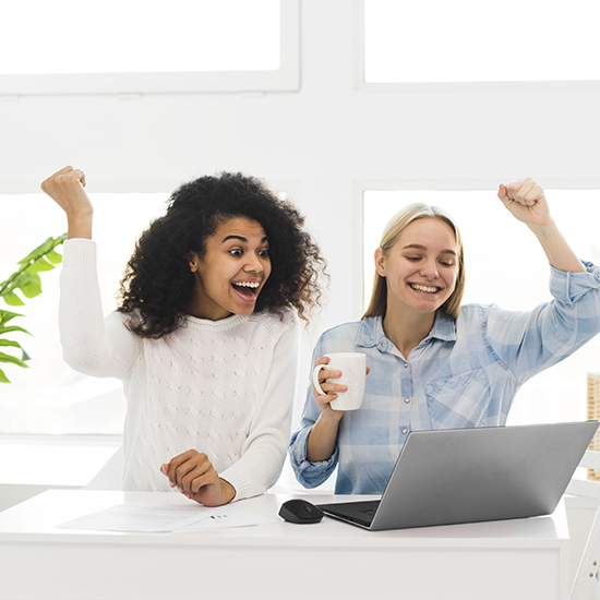 Two women are sitting at a desk in a bright room, celebrating and smiling while looking at a laptop screen. The woman on the left has curly hair and wears a white sweater. The woman on the right, with straight blonde hair, holds a coffee mug and wears a plaid shirt.