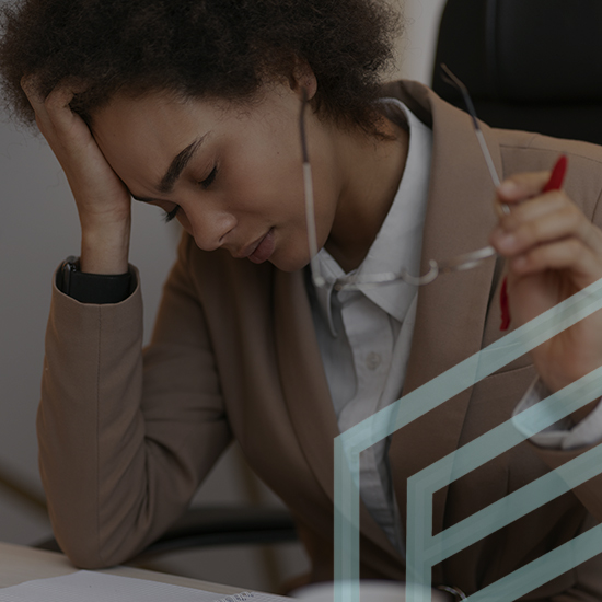 A person with curly hair is sitting at a desk with a hand on their forehead, eyes closed, and holding glasses in the other hand. They are wearing a beige blazer and white shirt, and appear to be stressed or fatigued. A piece of paper and red pen are on the desk.