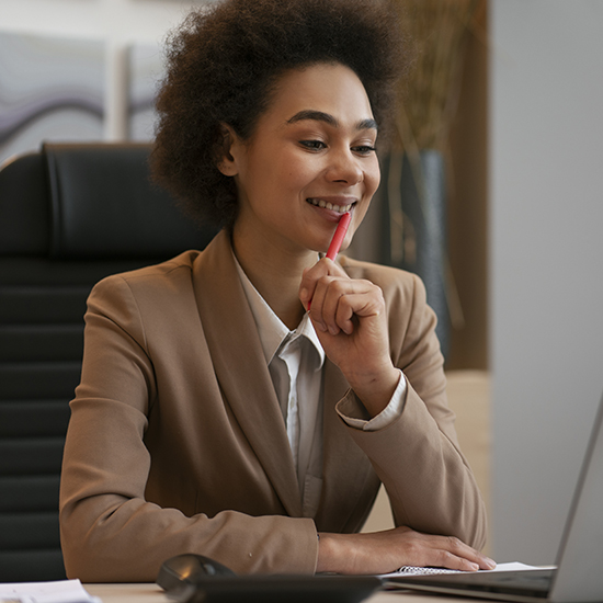 A woman with curly hair, wearing a beige blazer and a white shirt, sits at a desk in an office environment. She is smiling and holding a red pen near her mouth while looking at a laptop screen. There are papers and a phone on the desk.