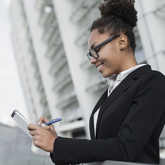 A person wearing a black blazer and white shirt is standing outside a modern building. They hold a smartphone in one hand and a pen in the other, smiling and looking at the device. The background shows an angular structure with balconies.