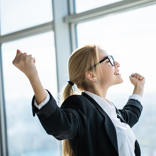 A woman with blonde hair tied back is wearing glasses, a white blouse, and a black blazer. She is smiling and looking up, with both arms raised in celebration. She is standing in front of large windows with natural light streaming in.