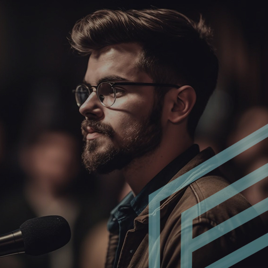 A man with glasses and a beard stands in front of a microphone, appearing to be engaged in public speaking or performing. The background is blurred, emphasizing the focus on the man, and there are geometric lines partially overlaying the image.