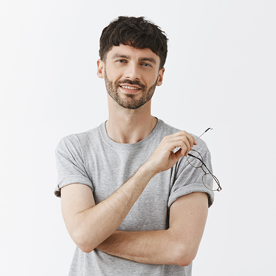 A smiling man with short, wavy dark hair and a beard is standing against a plain white background. He is holding a pair of eyeglasses in his right hand and has his left arm crossed over his chest. He is dressed in a light grey rolled-up sleeved T-shirt.