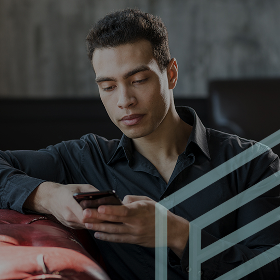 A young man with short dark hair is sitting on a red couch, looking at his smartphone with a focused expression. He is wearing a black shirt and resting one arm on the back of the couch. The background is a dimly lit, modern interior.