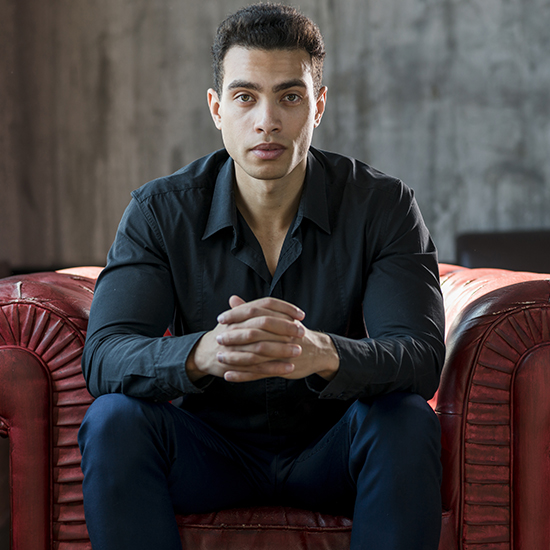 A man with short, dark hair and a serious expression is sitting on a red leather couch. He is wearing a black shirt and dark pants, with his hands clasped together in front of him. The background is a plain, textured wall.