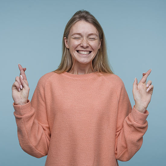 A woman with long blonde hair, wearing a peach-colored sweater, stands against a light blue background. She is smiling broadly with her eyes closed and both hands raised, fingers crossed on each hand.