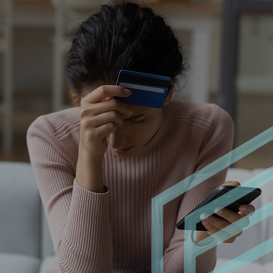 A woman sits looking stressed, holding a credit card to her forehead with one hand and a mobile phone in the other. She is wearing a pink sweater and her hair is tied back. The background shows blurred furniture, suggesting she is in a home setting.