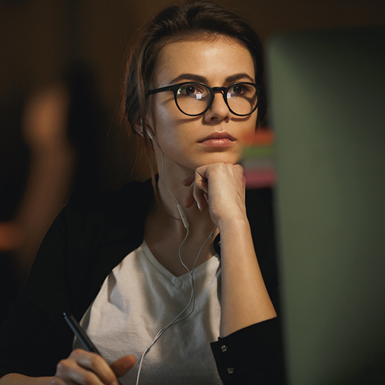 A person with glasses is sitting and looking at a computer screen. They are wearing a white shirt and dark sweater, with earphones in their ears. One hand is resting on their chin, while the other holds a pen. The background is dimly lit.