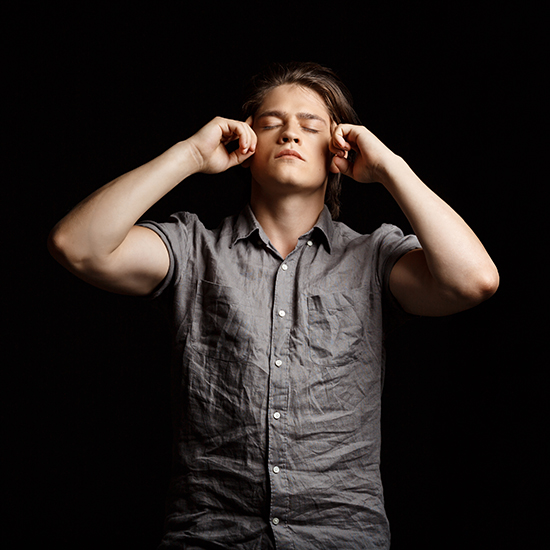 A young man with medium-length hair, wearing a short-sleeved gray shirt, stands against a black background. He has a calm expression and his eyes are closed. Both of his hands are raised, with his fingers pressing against his temples.