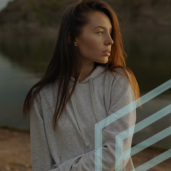 A woman with long brown hair wearing a light gray hoodie stands near a body of water, looking to her left. The background is a rocky shoreline, and there is a graphic overlay of intersecting light blue lines at the bottom right of the image.