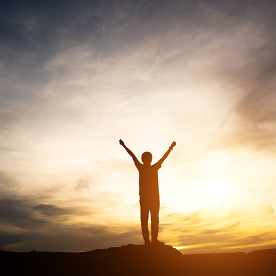 A person stands on a hilltop with arms raised triumphantly against a background of a dramatic sunset with vibrant orange, yellow, and purple hues in the sky. The silhouette is highlighted by the setting sun.