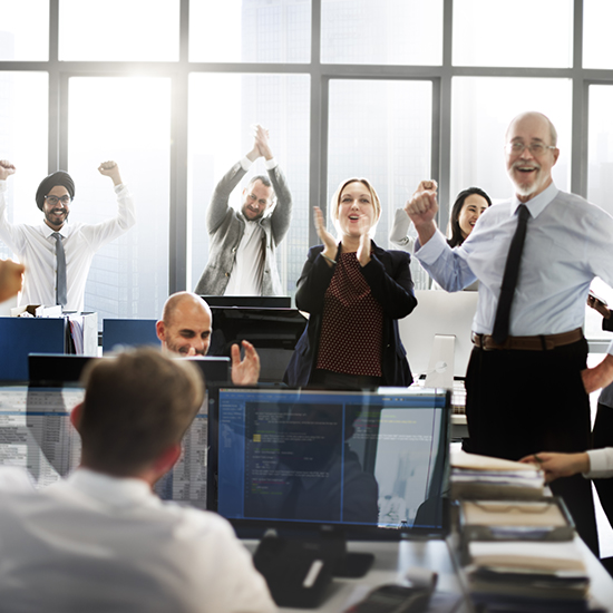 An office with large windows shows a diverse group of people celebrating enthusiastically. Some are clapping, others are cheering or raising their fists. Several monitors and desks with office supplies are visible in the foreground.