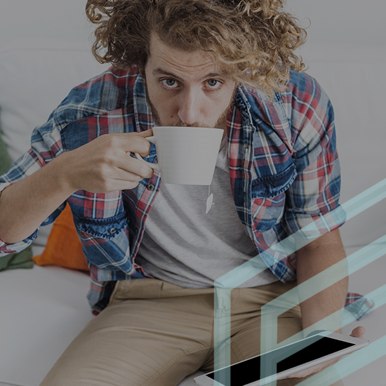 A curly-haired man in a plaid shirt and khaki pants sits on a white couch, sipping from a white mug while holding a tablet. There is a decorative glass structure in the foreground.