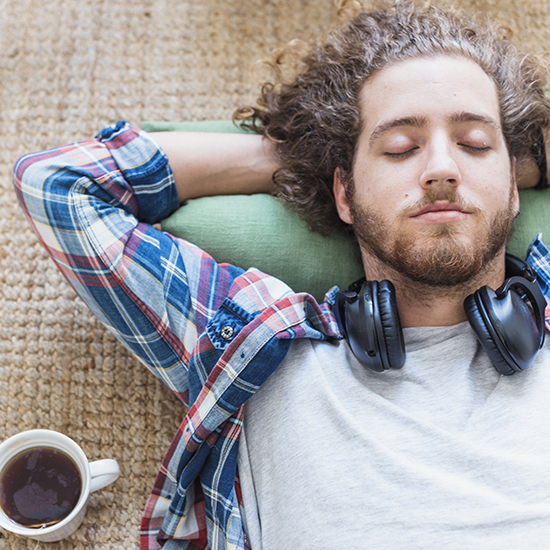 A person with curly hair is lying on a green pillow with headphones around their neck and eyes closed, appearing relaxed. They are wearing a plaid shirt over a grey t-shirt, and a cup of tea is placed on a woven rug next to them.