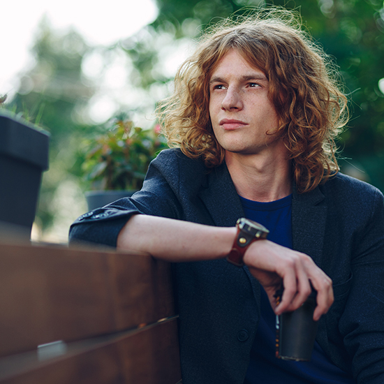 A young person with curly, shoulder-length hair and wearing a blazer sits leaning against a wooden bench, holding a cup. They look off into the distance with a thoughtful expression. The background is blurred with greenery and potted plants.