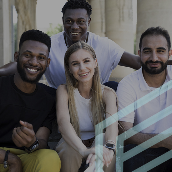 A group of four people, three men and one woman, sitting on steps and smiling at the camera. They appear to be outside near a building with tall columns. The image has a partially transparent geometric overlay.