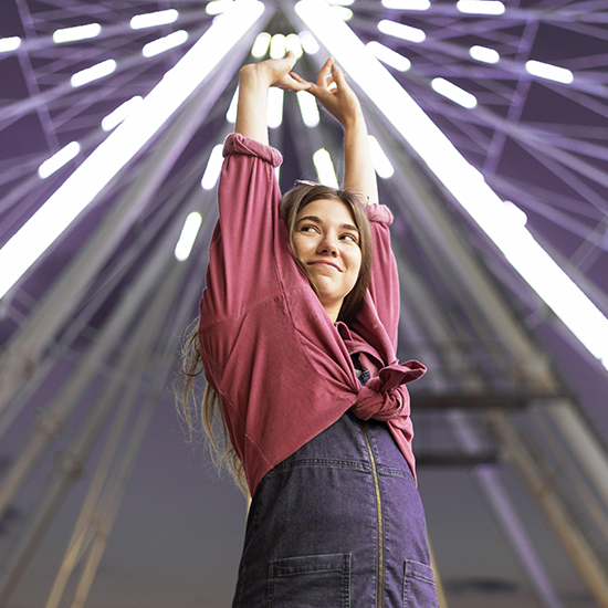 A young woman in a pink shirt and denim overalls smiles and stretches her arms above her head. She stands in front of a brightly lit Ferris wheel at night, creating a dynamic, joyful atmosphere.