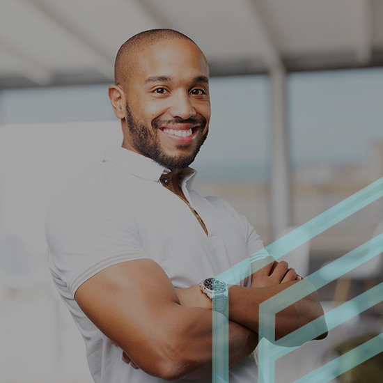 A man with a beard and mustache is smiling and standing with his arms crossed. He is wearing a white polo shirt and a wristwatch. The background appears to be a modern, indoor setting with large windows. Geometric shapes are overlaid on the right side of the image.