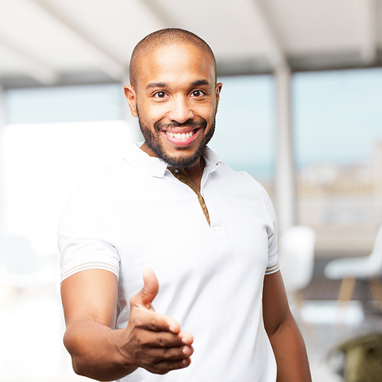 A smiling man extends his hand for a handshake. He is wearing a white polo shirt and stands in a bright room with large windows in the background. The setting appears to be modern and well-lit.