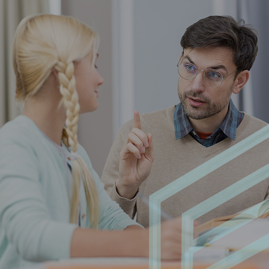 A man with glasses is talking to a young girl with blonde hair in a braid. They are seated at a table, and the man is gesturing with his finger as if explaining or emphasizing a point. The setting appears to be an indoor environment, possibly an office or classroom.