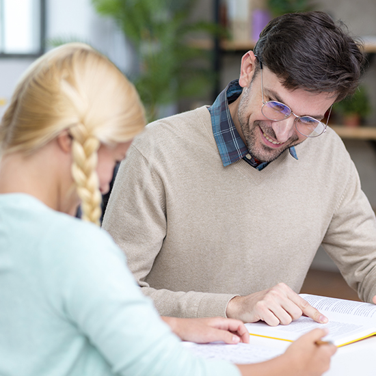 A man with glasses and a sweater is smiling and pointing at a book while sitting across from a woman with blonde braided hair. They are at a table in a bright room with plants in the background, focusing on the book.