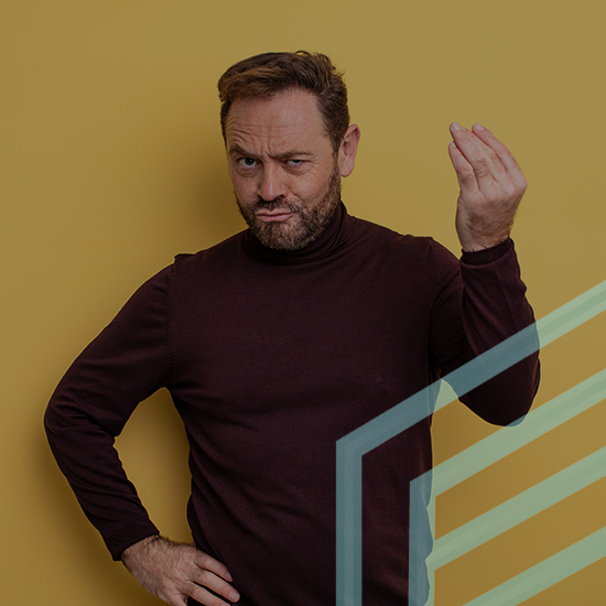 A man with a beard and short hair, wearing a dark maroon turtleneck, stands against a mustard-yellow background. He raises one hand, fingers pinched together, while his other hand is on his hip. He has an expressive look on his face.