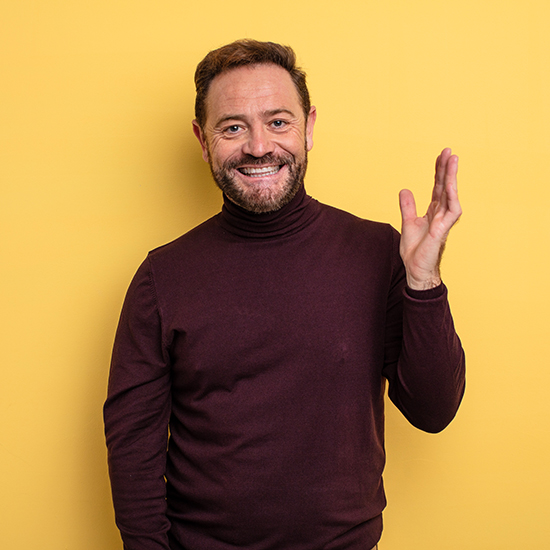 A man with a beard and short hair is smiling and waving at the camera. He is wearing a maroon turtleneck sweater and stands against a solid yellow background.