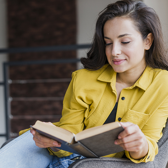 A person with long dark hair, wearing a yellow shirt, sits on a couch reading a book. They appear relaxed and focused, with a slight smile on their face and a background that shows part of a room with a metal railing.