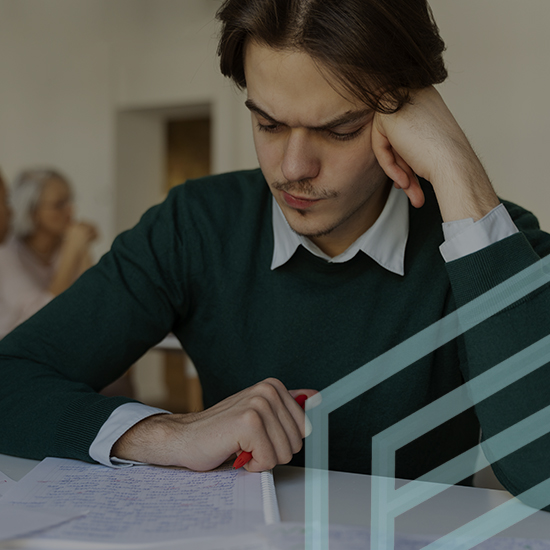 A young man with brown hair is sitting at a desk, looking intently at a sheet of paper while holding a red pen. He is wearing a green sweater over a white collared shirt. Two blurry figures are visible in the background. A geometric design overlays part of the image.