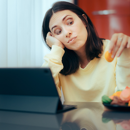 A woman with shoulder-length dark hair, wearing a light yellow sweater, sits at a table looking at a tablet with a bored expression. She holds a piece of fruit in one hand and rests her head on the other, with a bowl of fruit in front of her.