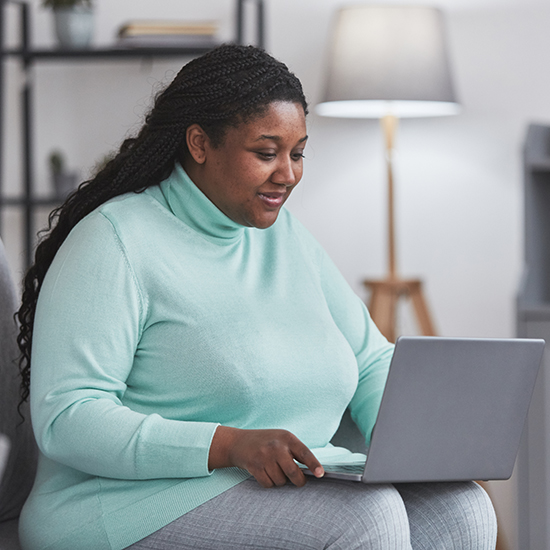 A woman with long braided hair wearing a light blue turtleneck sweater is sitting on a couch, smiling and looking at a laptop in her lap. A lamp and a bookshelf are visible in the background.