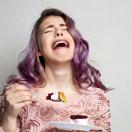 A woman with purple hair and a pink floral top holds a plate with a piece of cake and a fork, looking distressed and appearing to cry. The creamy cake has a layer of fruit topping. The background is plain and light-colored.