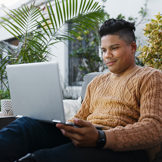 A person with short, dark hair sits outside on a cushioned seat, holding an open laptop on their lap. They are wearing a tan, cable-knit sweater and dark pants. Tropical plants surround them, creating a serene atmosphere. The person appears focused on the screen.