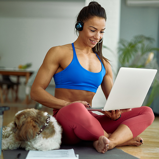 A woman in a blue sports bra and red leggings sits on a mat inside a living room, using a laptop. She has headphones on and is smiling. A small dog lies beside her, looking up. There are papers on the floor nearby.