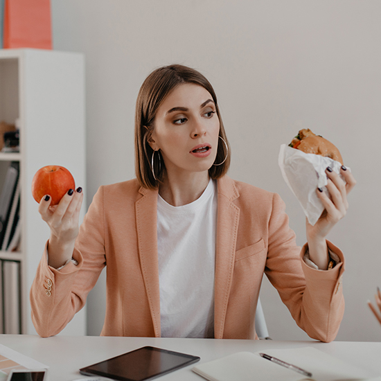 A woman with medium length hair, wearing a peach blazer, sits at a desk holding an apple in one hand and a wrapped burger in the other. She appears to be contemplating her choice between the two foods. The desk has notebooks, a tablet, and office supplies.