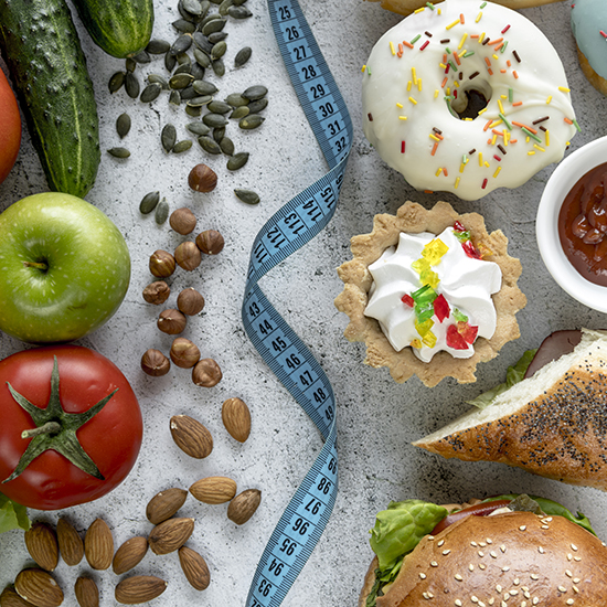 A variety of foods, including apples, tomatoes, a cucumber, a doughnut, a tart, seeds, nuts, a sandwich, and a burger are arranged on a table. A blue measuring tape is draped across the middle of the image.