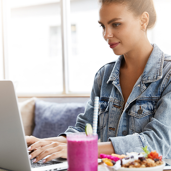 A woman with a ponytail works on a laptop at a table near a window. She is wearing a denim jacket and has a focused expression. There is a pink smoothie in a mason jar with a straw and a bowl of granola with fruit on the table in front of her.