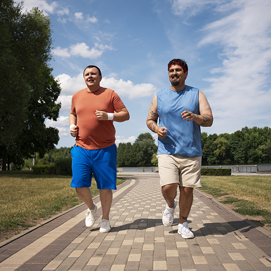 Two men are jogging on a paved path in a park on a sunny day. One wears a red-orange shirt and blue shorts, while the other wears a blue sleeveless shirt and beige shorts. Both are smiling and surrounded by greenery and trees under a partly cloudy sky.