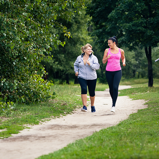 Two women are jogging along a winding dirt path in a park. One is wearing a gray jacket and black leggings, while the other is in a pink tank top and black leggings. They are smiling and engaged in conversation, surrounded by green trees and grass.