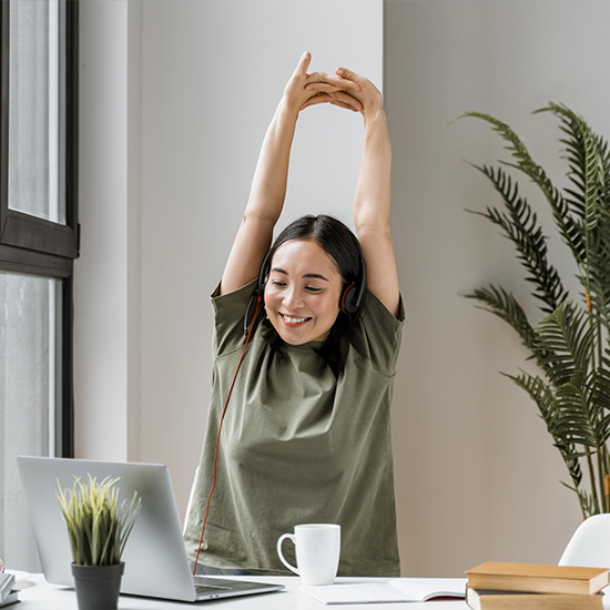 A woman wearing a green shirt and headphones is sitting at a desk with a laptop, a cup, and books. She is stretching her arms above her head and smiling. There is a potted plant on the desk and another larger potted plant in the background near a window.