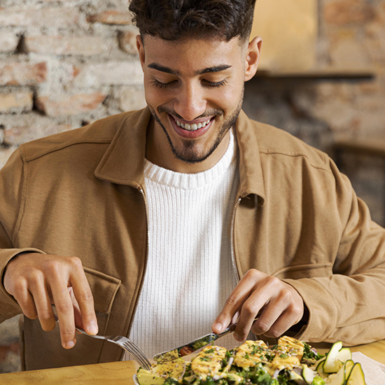 A man with short curly hair and a trimmed beard is seated at a wooden table, smiling as he cuts into a dish with vegetables and sauce. He is wearing a tan jacket over a white sweater. The background features a rustic brick wall.