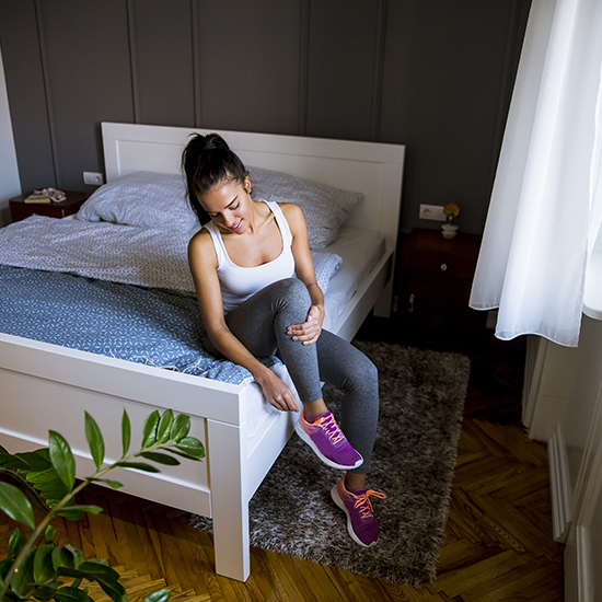 A woman in athletic wear sits on the edge of a bed, with one leg lifted and hands holding her ankle. She appears to be in a bedroom with a wooden floor, gray wall, and a large window with curtains. There is a plant in the foreground and bedside tables next to the bed.