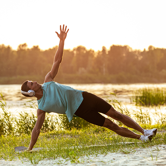 A person wearing headphones performs a side plank exercise outdoors by a river at sunset, balancing on one hand and raising the other arm while extending their legs. The scene includes greenery, water, and a forested backdrop.