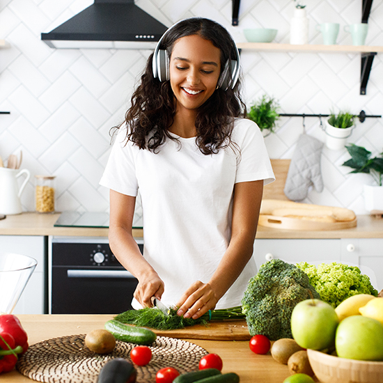 A woman with long curly hair, wearing a white t-shirt and headphones, smiles while cutting vegetables in a bright, modern kitchen. Fresh produce including tomatoes, apples, broccoli, and bell peppers are spread out on the counter.