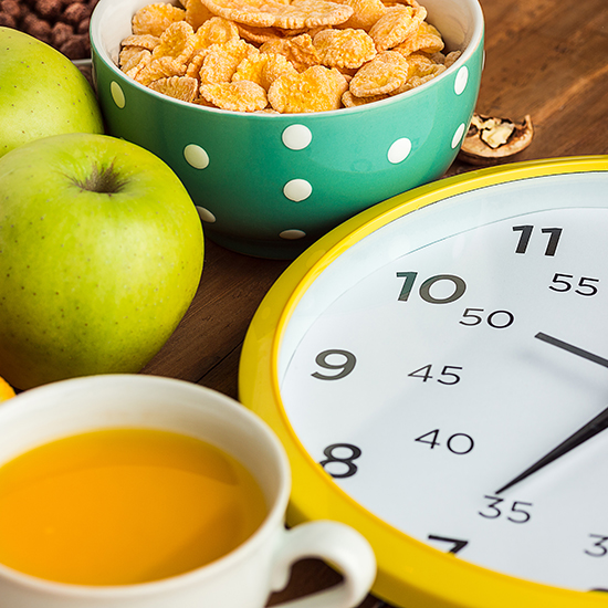 A breakfast scene featuring a bowl of cereal in a green polka dot bowl, a cup of orange juice, two green apples, and a yellow-rimmed clock displaying approximately 9:05.