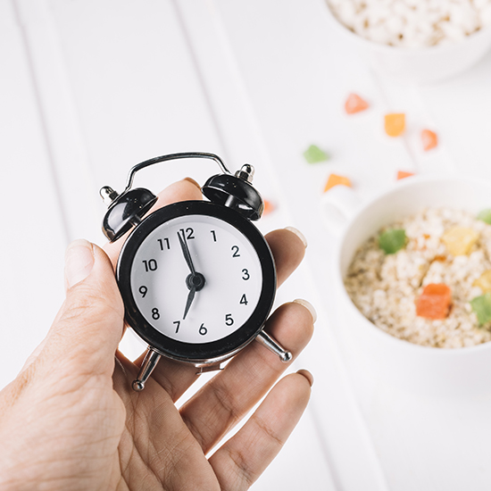 A hand holds a small black alarm clock showing 12:00. In the blurred background, there are white bowls filled with grains and colorful diced vegetables placed on a white surface. The scene suggests a focus on time and food.