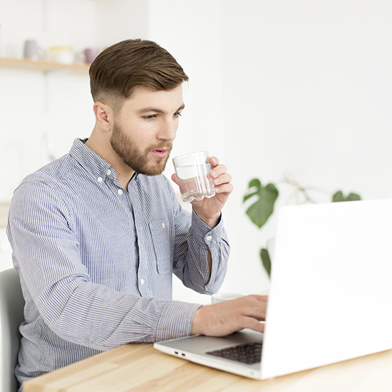 A man in a blue striped shirt sits at a table, working on a laptop. He is holding a glass of water and appears to be focused on the screen. The background features a small plant and shelves with a few items placed neatly on them.