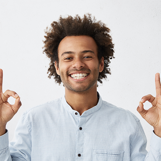 A man with curly hair is smiling broadly and looking at the camera. He is wearing a light blue button-up shirt and has both hands raised, making an "OK" gesture with his thumb and index fingers. The background is plain white.