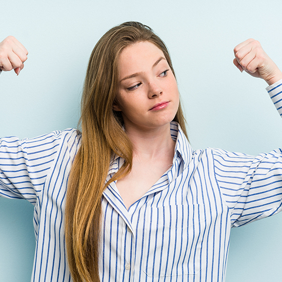 A young woman with long hair, wearing a striped shirt, poses against a light blue background. She looks confident as she flexes both arms, showing off her biceps and giving a slight smile.